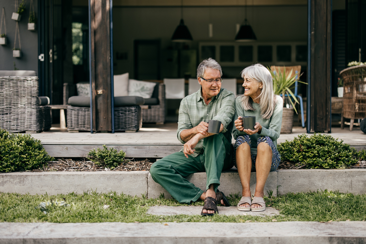 Seated on the outdoor steps of their stylish home, a mature couple enjoys a peaceful morning with their favorite drinks.